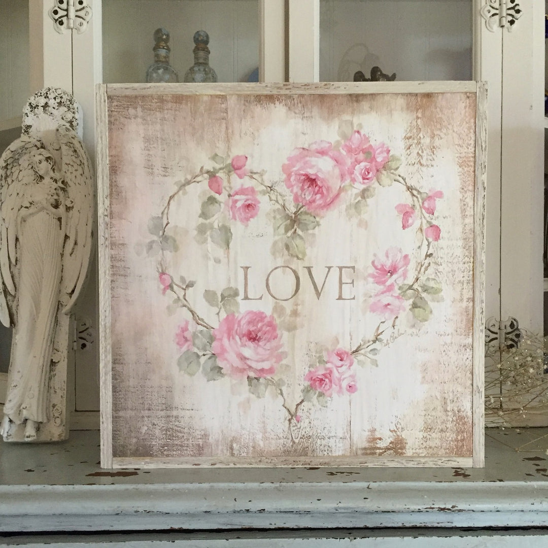 A heart of pink roses, large and small including buds with greenery surrounding the words "Love" . Background is a distressed off white with lots of antiqueing, all framed in a cream color barnwood frame.