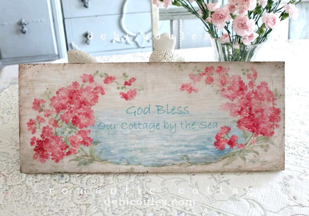 Beautiful branches of pink and red crape myrtle frame a glimse of the blue sea. Background is off white with a distressed look. A simple line says "Good bless our cottage by the sea".