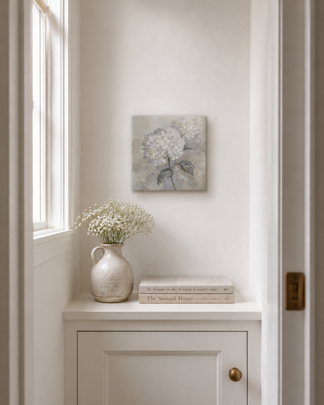Nook with a vase of flowers, books, and a small square hyprangea painting on a white wall.