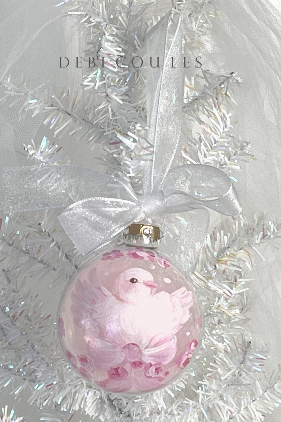 Shabby-chic, glass Christmas ornament with a dove and pink bow design hanging on a white tinsel background.