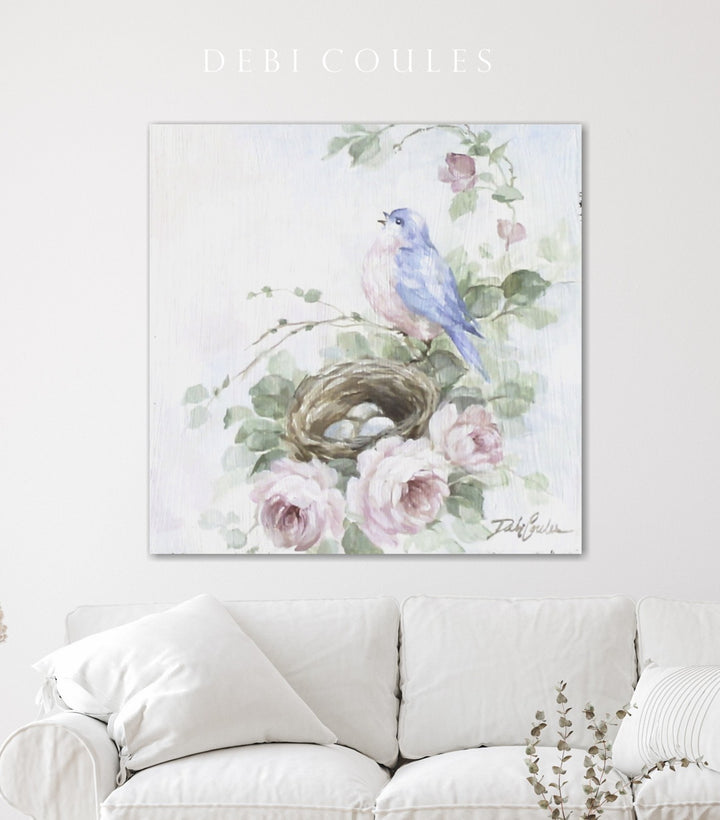 A canvas giclée print featuring a bluebird perched on a branch with a nest, surrounded by pink roses, displayed above a white sofa.