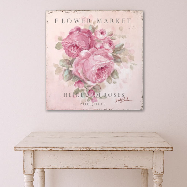 A large cottage sign of pink heirloom roses hangs above a distressed wood table painted white.