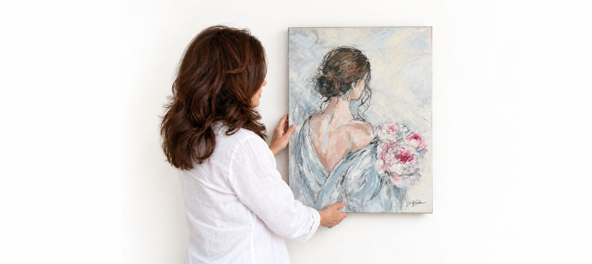 Artist Debi Coules hanging her new original painting of a woman holding a bouquet of pink peonies
