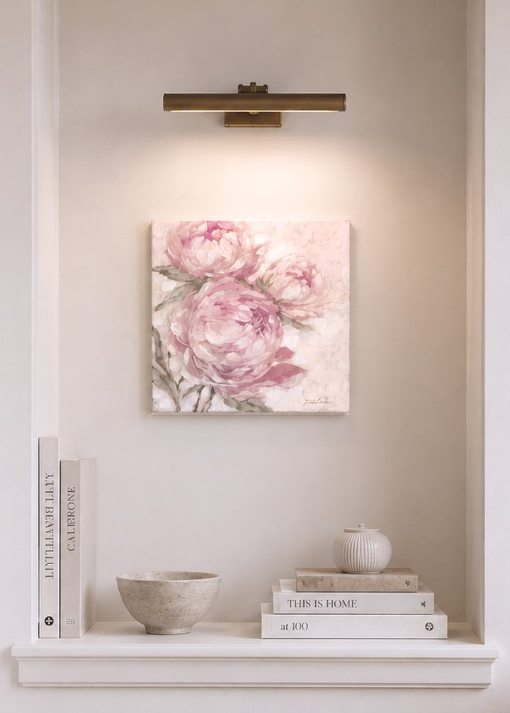Floral pink painting with overhead light on a wall above a shelf with books and decor items.