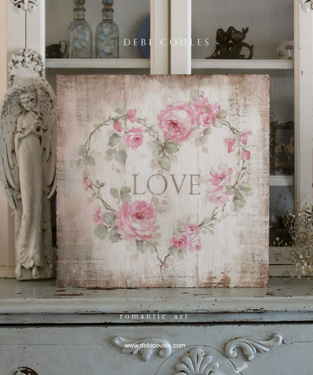 A heart of pink roses, large and small including buds with greenery surrounding the words "Love" . Background is a distressed off white with lots of antiqueing
